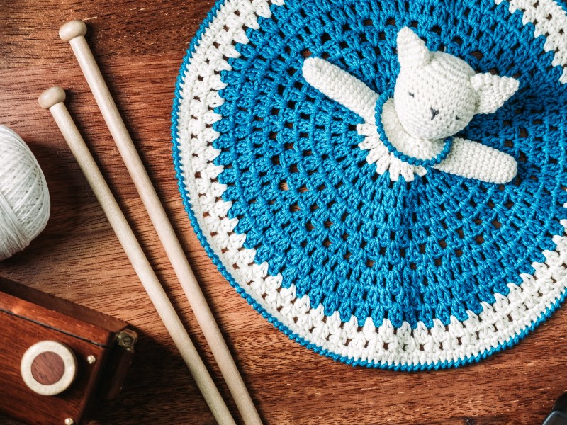 A crocheted teddy bear and knitting needles on a wooden table.