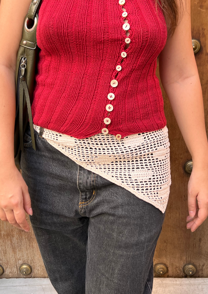 A person wearing a red sleeveless ribbed top with diagonal buttons, dark jeans, a cream crochet hip wrap, and carrying a green bag.