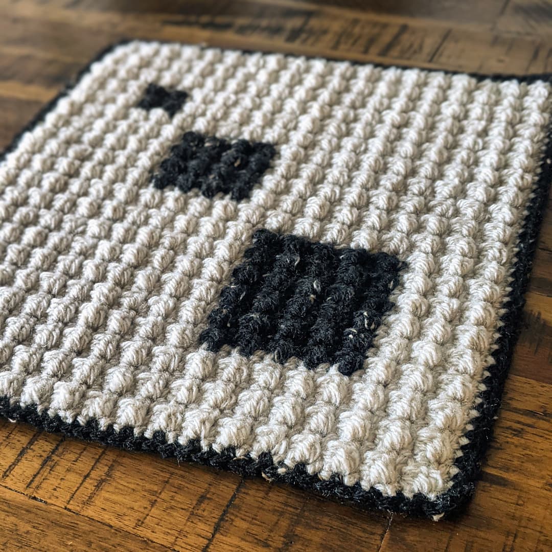 A crocheted square mat with a cream background, two black squares, and a black border, placed on a wooden surface.